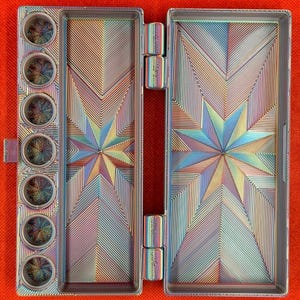 May include: A rectangular metal watercolor palette with an iridescent finish. The interior features seven circular paint wells on the left and two large rectangular mixing areas, all decorated with a geometric starburst pattern of fine lines.
