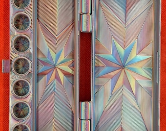 May include: A rectangular metal watercolor palette with an iridescent finish. The interior features seven circular paint wells on the left and two large rectangular mixing areas, all decorated with a geometric starburst pattern of fine lines.