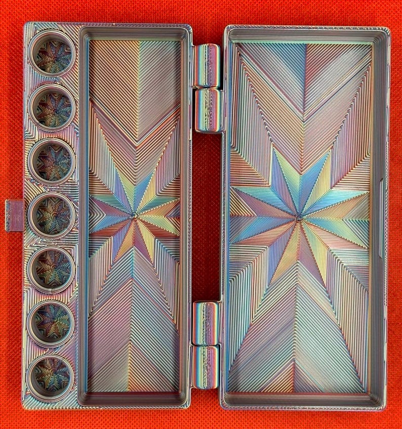 May include: A rectangular metal watercolor palette with an iridescent finish. The interior features seven circular paint wells on the left and two large rectangular mixing areas, all decorated with a geometric starburst pattern of fine lines.