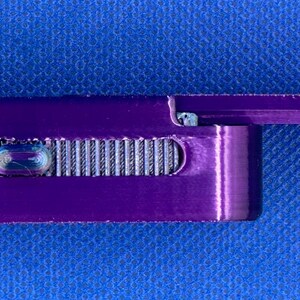May include: A rectangular purple metal smoking pipe featuring a sliding lid mechanism and a textured silver-coloured grip slider on the side. The device has a smooth, metallic finish and a compact, elongated shape with rounded edges.