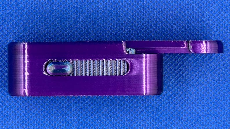 May include: A rectangular purple metal smoking pipe featuring a sliding lid mechanism and a textured silver-coloured grip slider on the side. The device has a smooth, metallic finish and a compact, elongated shape with rounded edges.