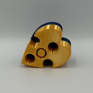 May include: A gold-colored, heart-shaped object with multiple circular openings. The top edge features a dark blue, velvety material. The object appears to be 3D-printed and has a textured surface. The design includes a central circular feature.