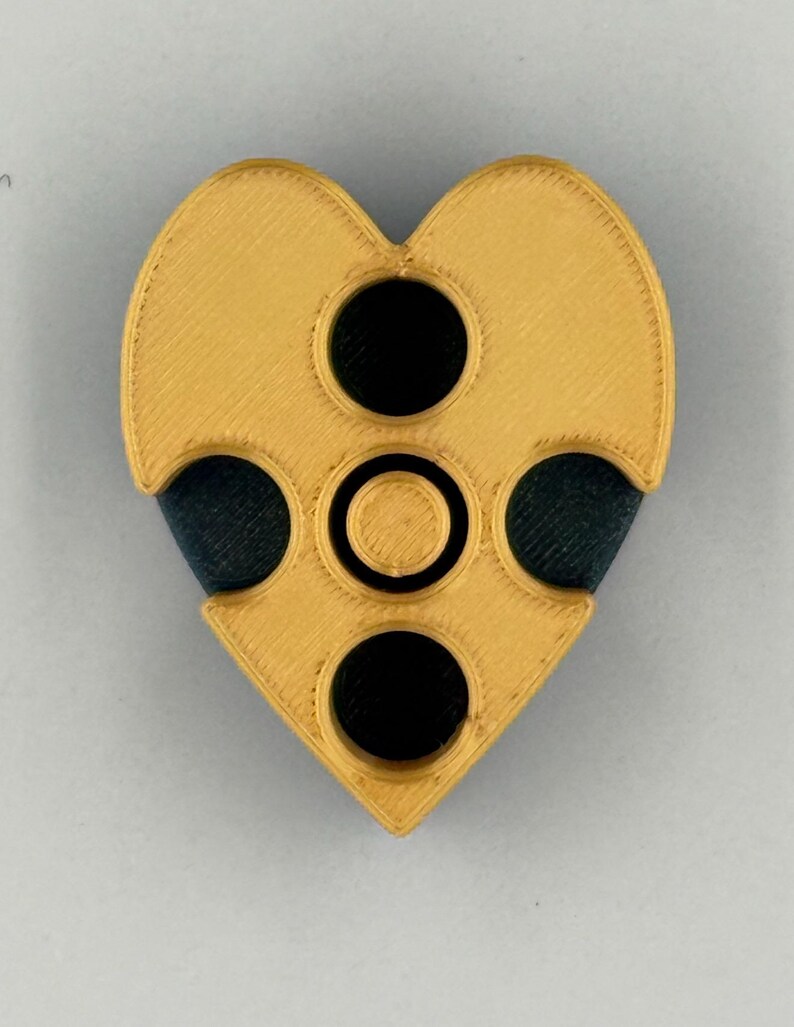 May include: A heart-shaped object with a gold-colored exterior and three black circular insets. The object has a textured surface and a geometric design, with a smaller circle in the center.