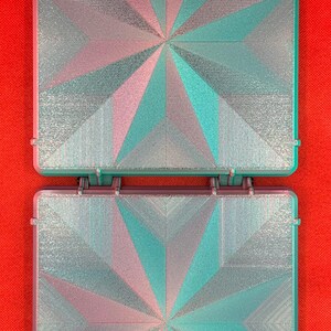 May include: Two square, hinged cases featuring a geometric star pattern on the lids. The surface has a textured, iridescent finish that shifts between shades of metallic silver, teal, and soft pink, creating a multi-faceted appearance.