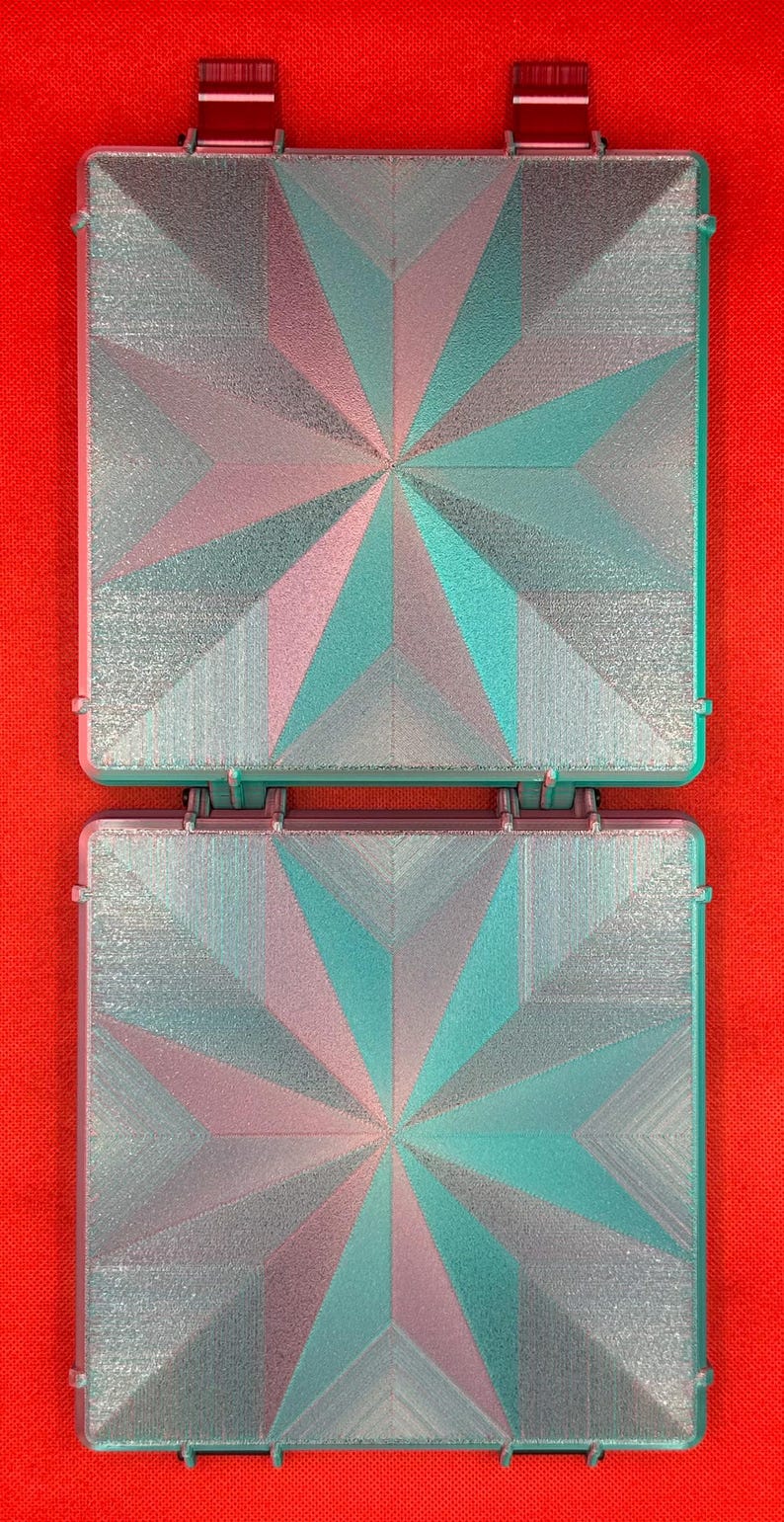 May include: Two square, hinged cases featuring a geometric star pattern on the lids. The surface has a textured, iridescent finish that shifts between shades of metallic silver, teal, and soft pink, creating a multi-faceted appearance.