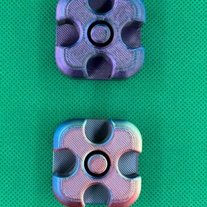 May include: Two square-shaped fidget spinners with rounded corners and a central circular bearing. Each piece features a gradient finish shifting between metallic purple, blue, and pink tones, with visible 3D printed layer lines.