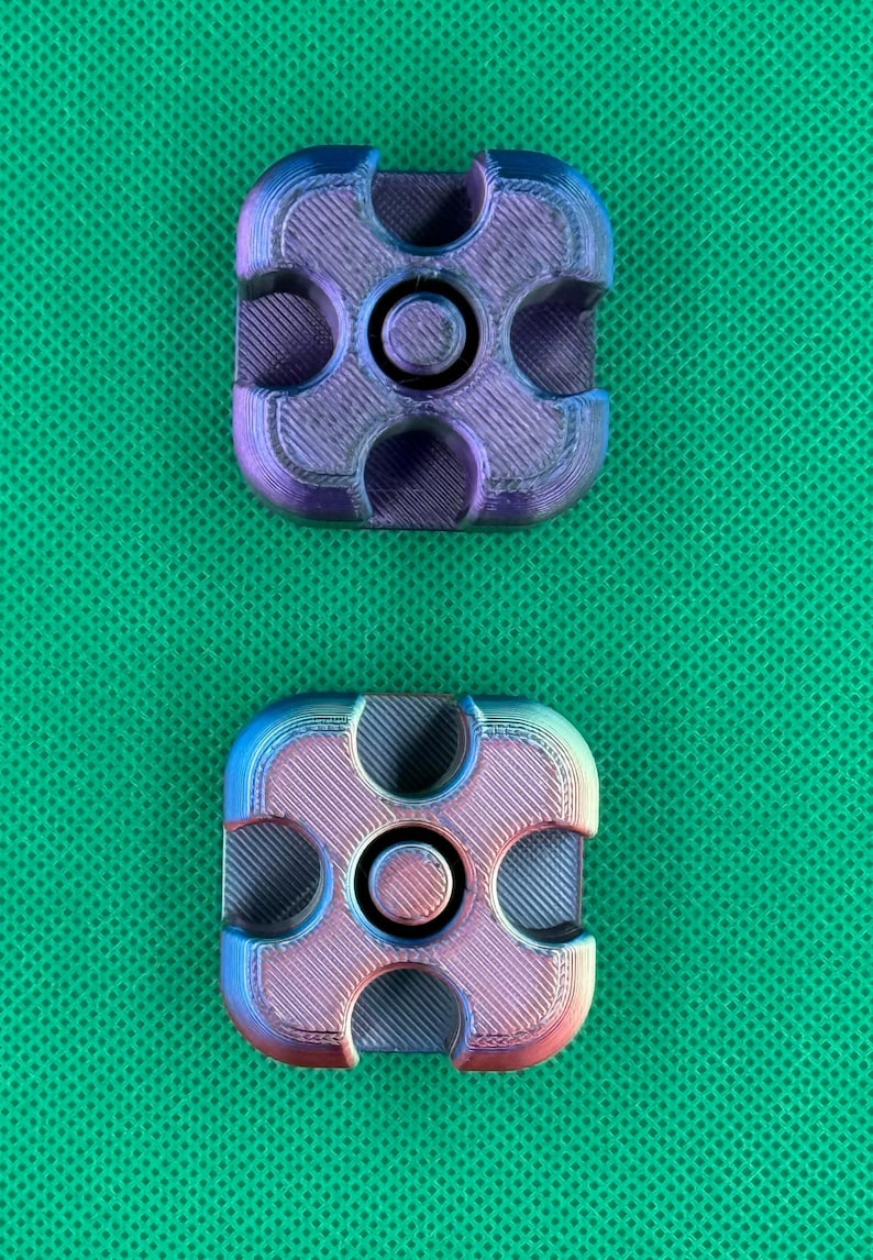 May include: Two square-shaped fidget spinners with rounded corners and a central circular bearing. Each piece features a gradient finish shifting between metallic purple, blue, and pink tones, with visible 3D printed layer lines.