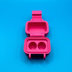 May include: A bright pink, rectangular, 3D-printed storage case with a hinged lid. The interior of the base features two circular recessed compartments, while the lid interior is hollow. The case includes a black latch mechanism.