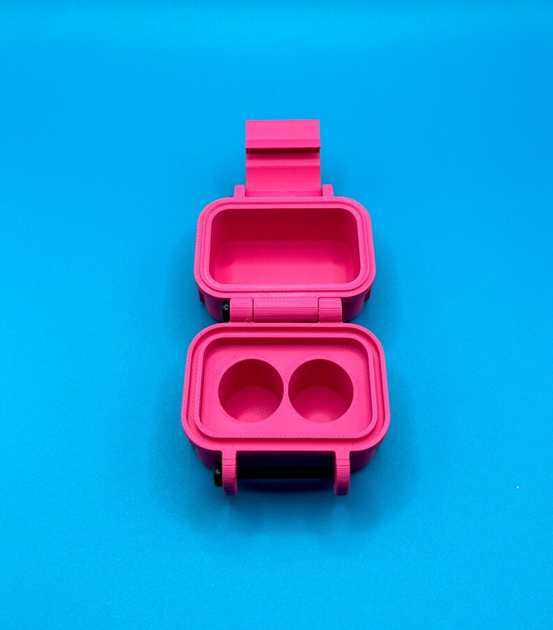 May include: A bright pink, rectangular, 3D-printed storage case with a hinged lid. The interior of the base features two circular recessed compartments, while the lid interior is hollow. The case includes a black latch mechanism.