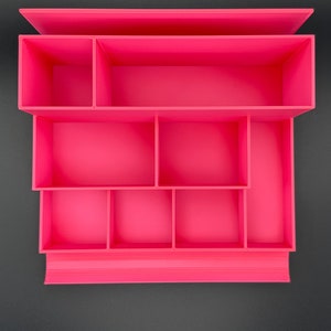 May include: A bright pink desk organizer featuring a tiered design with multiple rectangular and square compartments of varying sizes. The structure is open-topped, providing organized storage sections for small items and office supplies.