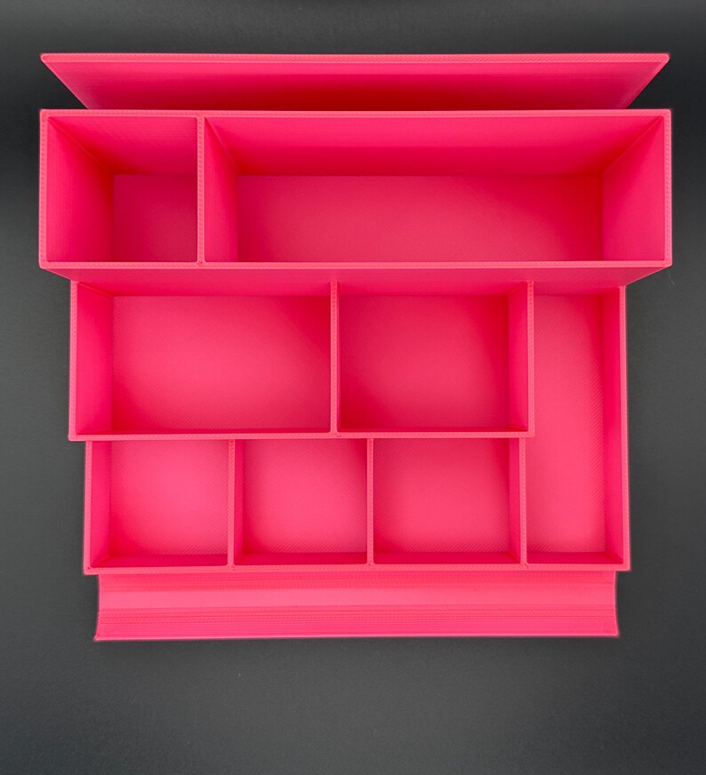 May include: A bright pink desk organizer featuring a tiered design with multiple rectangular and square compartments of varying sizes. The structure is open-topped, providing organized storage sections for small items and office supplies.