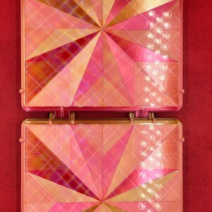 May include: Two square-shaped, pink and gold, decorative tiles with a geometric pattern. The tiles are made of a shiny, iridescent material.