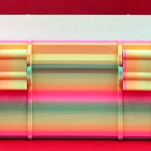 Puede incluir: Una caja de almacenamiento de pl&aacute;stico con colores de arco&iacute;ris, con dos cierres y una tapa texturizada. La caja tiene un dise&ntilde;o de rayas multicolores con tonos rosas, naranjas, amarillos, verdes y azules.