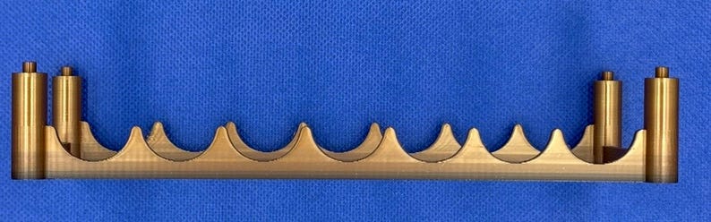 K&ouml;nnte beinhalten: Ein goldfarbener Metallst&auml;nder mit einer Reihe von geschwungenen Vertiefungen, die f&uuml;r die Aufnahme mehrerer Gegenst&auml;nde ausgelegt sind. Der St&auml;nder hat an jedem Ende zwei vertikale Pfosten.