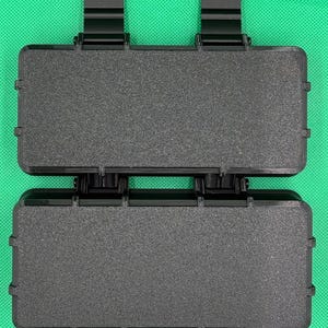 Puede incluir: Un estuche de almacenamiento rectangular negro con una superficie texturizada y dos bisagras negras prominentes en el borde superior. El estuche presenta una construcci&oacute;n r&iacute;gida y duradera con un mecanismo de cierre seguro.