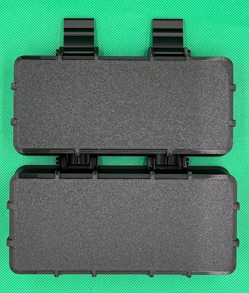 Puede incluir: Un estuche de almacenamiento rectangular negro con una superficie texturizada y dos bisagras negras prominentes en el borde superior. El estuche presenta una construcci&oacute;n r&iacute;gida y duradera con un mecanismo de cierre seguro.