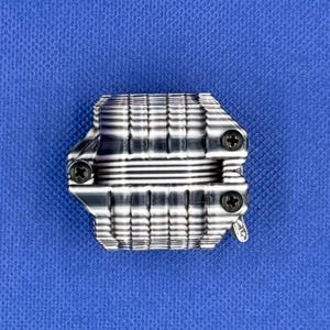 K&ouml;nnte beinhalten: Eine metallische, rechteckige Komponente mit einer Reihe horizontaler, gerippter Lamellen und drei sichtbaren schwarzen Kreuzschlitzschrauben. Das Objekt hat ein strukturiertes, industrielles Aussehen mit polierter Oberfl&auml;che und einer kleinen Metalllasche an der Seite.