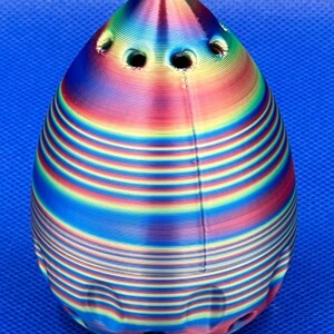 May include: A colorful, iridescent, 3D printed object shaped like a teardrop with multiple holes. The object has a striped pattern and a smooth, glossy finish.