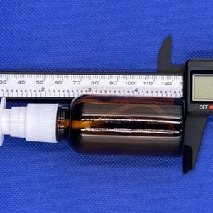 May include: A brown glass bottle with a white plastic nasal spray pump nozzle. The bottle is positioned against a digital caliper showing a measurement of 4.96 inches, indicating the total length of the bottle and nozzle assembly.