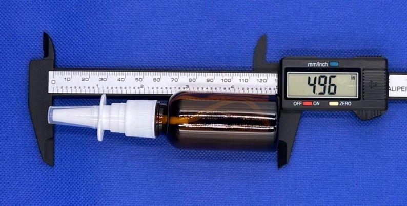 May include: A brown glass bottle with a white plastic nasal spray pump nozzle. The bottle is positioned against a digital caliper showing a measurement of 4.96 inches, indicating the total length of the bottle and nozzle assembly.