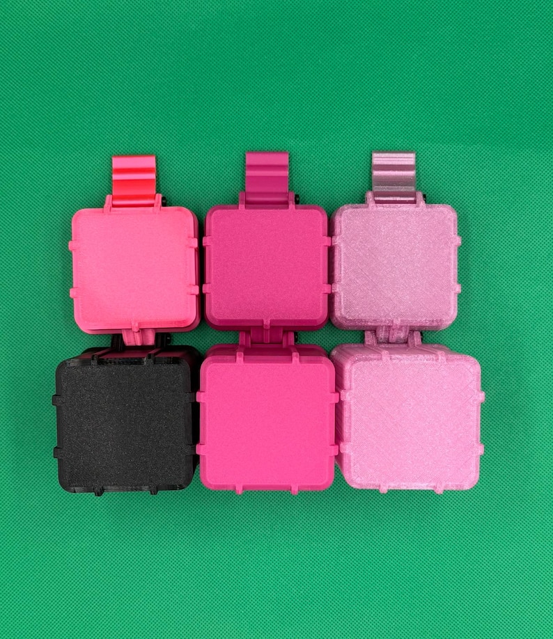 Puede incluir: Un conjunto de seis recipientes de almacenamiento cuadrados con bisagras, dispuestos en dos filas de tres. La fila superior presenta tonos rosa brillante, magenta y morado claro. La fila inferior incluye un recipiente negro, uno rosa intenso y uno rosa claro.