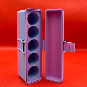 May include: A rectangular, light purple and blue gradient storage case with a hinged lid. The interior features a vertical insert with five circular cutouts for holding cylindrical items, secured by a small black metal hinge pin on the side.