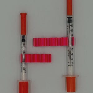 May include: Two insulin syringes with orange caps and plungers, and clear barrels marked with measurements. Two bright pink, ridged plastic pieces are positioned around the syringes. The syringes are on a white background.