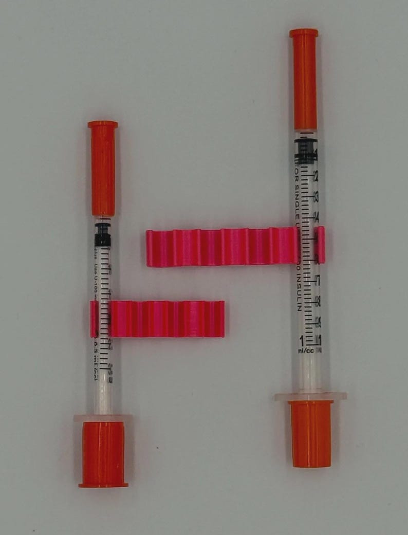May include: Two insulin syringes with orange caps and plungers, and clear barrels marked with measurements. Two bright pink, ridged plastic pieces are positioned around the syringes. The syringes are on a white background.