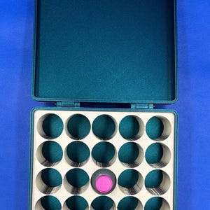 May include: A teal green storage case with a lid, containing 25 round compartments. One compartment has a pink and gray lid.