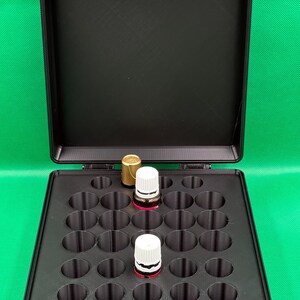 May include: A black rectangular storage case with a hinged lid, featuring a custom interior insert with twenty-five circular cutouts. Two small glass bottles with white screw-on caps are placed within the organised grid of the storage box.