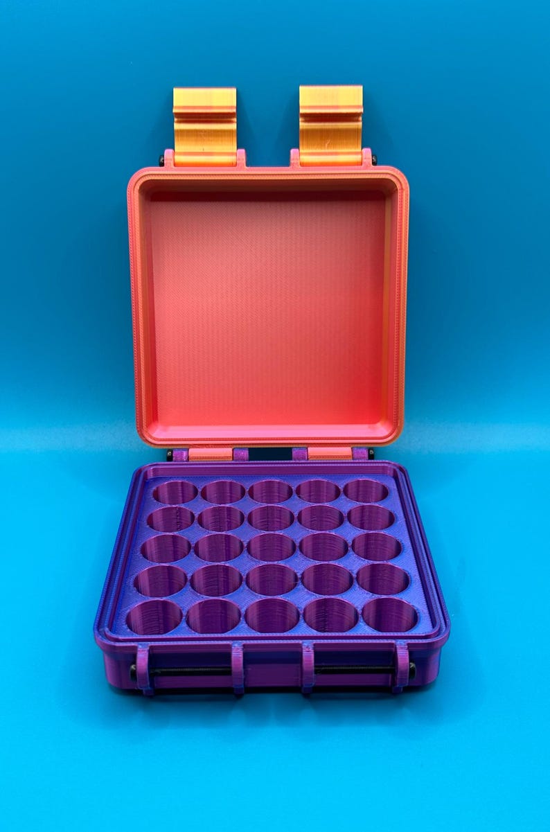May include: A rectangular storage case with an open lid, featuring a bright orange interior and exterior frame. The base contains a purple insert with twenty-five circular compartments arranged in a five-by-five grid for organized storage.