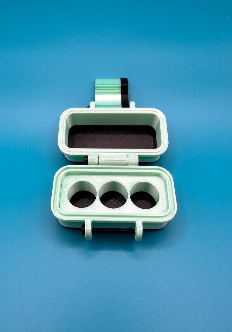 Puede incluir: Una caja rectangular con bisagras en color verde menta y negro con un cierre. La secci&oacute;n superior es un compartimento abierto, mientras que la secci&oacute;n inferior presenta tres aberturas circulares. Las superficies interiores son negras.