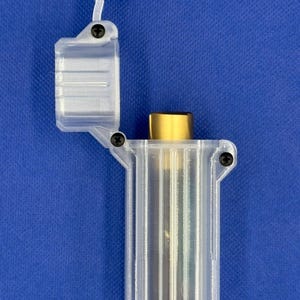 Puede incluir: Un estuche de pl&aacute;stico transparente y transl&uacute;cido impreso en 3D con tapa abatible, que contiene un objeto cil&iacute;ndrico de color dorado. El estuche est&aacute; asegurado con peque&ntilde;os tornillos negros en la bisagra y el lateral, la tapa est&aacute; abierta.