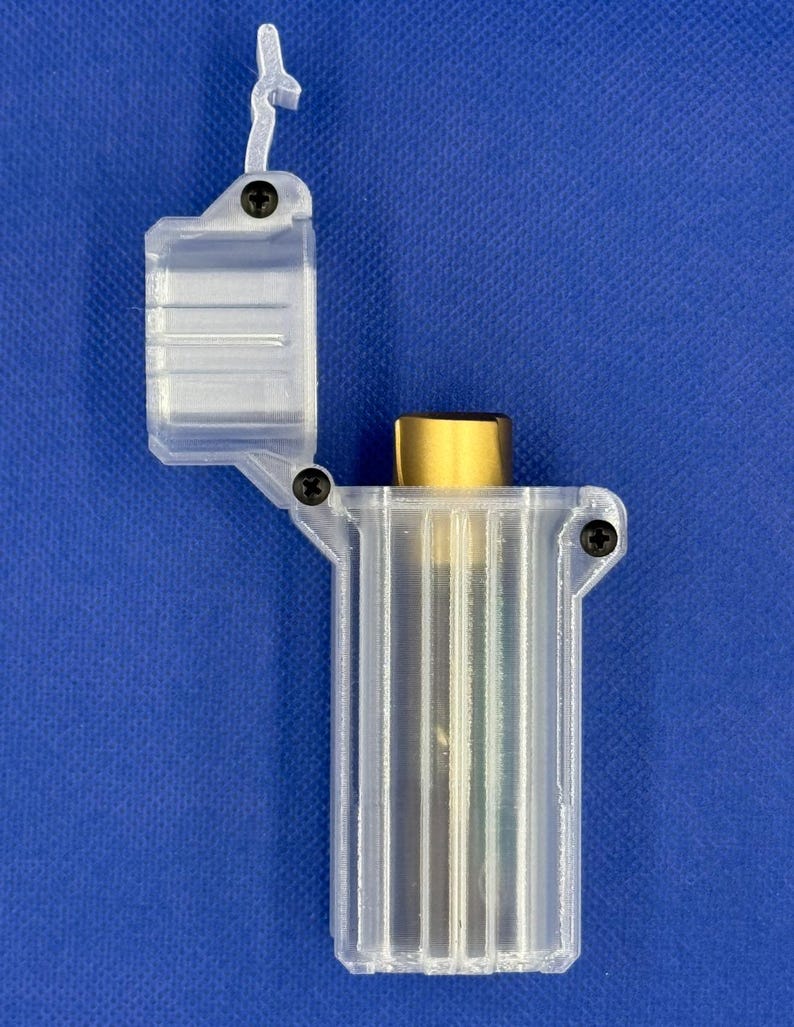 Puede incluir: Un estuche de pl&aacute;stico transparente y transl&uacute;cido impreso en 3D con tapa abatible, que contiene un objeto cil&iacute;ndrico de color dorado. El estuche est&aacute; asegurado con peque&ntilde;os tornillos negros en la bisagra y el lateral, la tapa est&aacute; abierta.