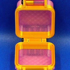 May include: A small, open, square-shaped storage container made of translucent pink plastic with a glittery finish. The container features a bright gold-colored frame, a hinged lid, and a black horizontal latch mechanism on the front.