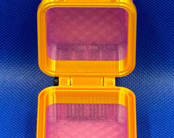 May include: A small, open, square-shaped storage container made of translucent pink plastic with a glittery finish. The container features a bright gold-colored frame, a hinged lid, and a black horizontal latch mechanism on the front.