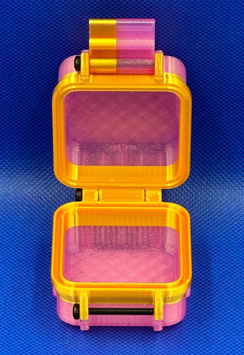 May include: A small, open, square-shaped storage container made of translucent pink plastic with a glittery finish. The container features a bright gold-colored frame, a hinged lid, and a black horizontal latch mechanism on the front.