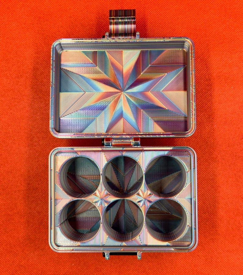 Pu&ograve; includere: Un contenitore metallico rettangolare con finitura iridescente, caratterizzato da un motivo a stella geometrica sul coperchio e sei scomparti circolari nella base. Le superfici interne presentano una trama lineare metallica multicolore.