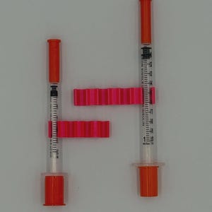 May include: Two insulin syringes with orange caps and plungers, and pink syringe holders. The syringes are clear with black measurement markings. The syringe holders are a bright pink color and have a ridged design.