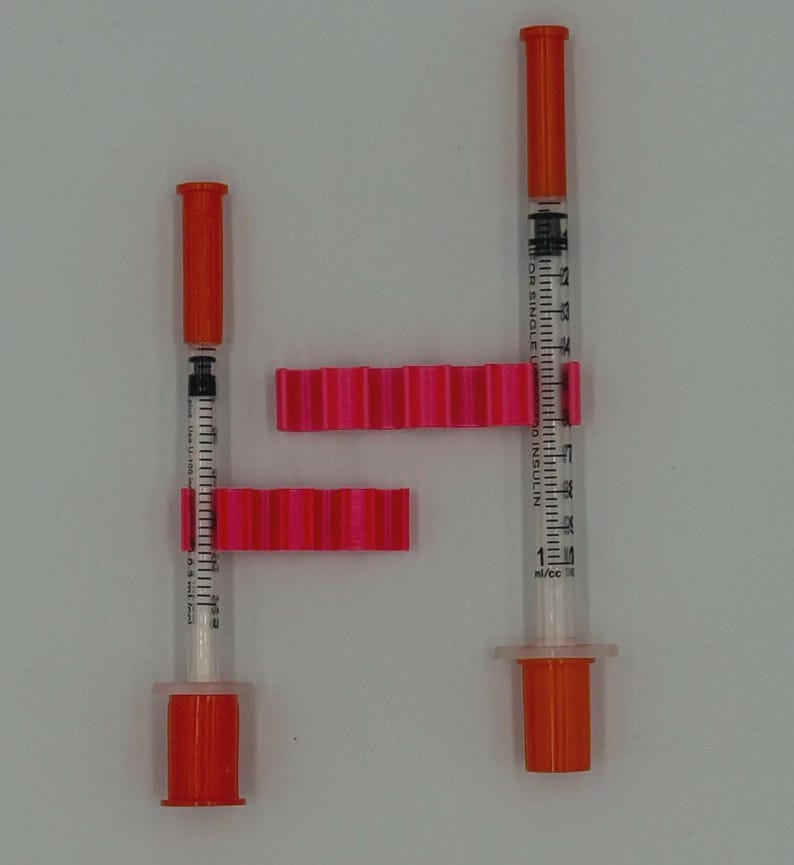 May include: Two insulin syringes with orange caps and plungers, and pink syringe holders. The syringes are clear with black measurement markings. The syringe holders are a bright pink color and have a ridged design.