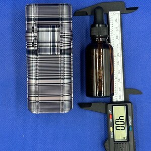 May include: A rectangular case with a black and white plaid pattern next to an amber glass dropper bottle. A black digital caliper measures the bottle, showing a reading of 4.00 inches on the display screen and a scale up to 100 mm.