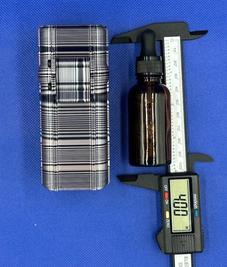 May include: A rectangular case with a black and white plaid pattern next to an amber glass dropper bottle. A black digital caliper measures the bottle, showing a reading of 4.00 inches on the display screen and a scale up to 100 mm.