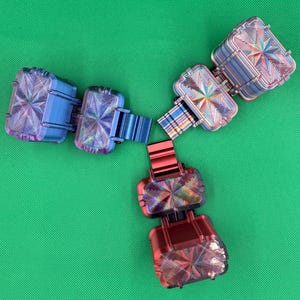Puede incluir: Tres juguetes antiestr&eacute;s articulados dispuestos en forma de Y. Cada juguete consta de dos bloques rectangulares conectados por una bisagra, con un acabado met&aacute;lico y un patr&oacute;n de estrella en la superficie. Uno es azul, otro rojo y otro iridiscente.