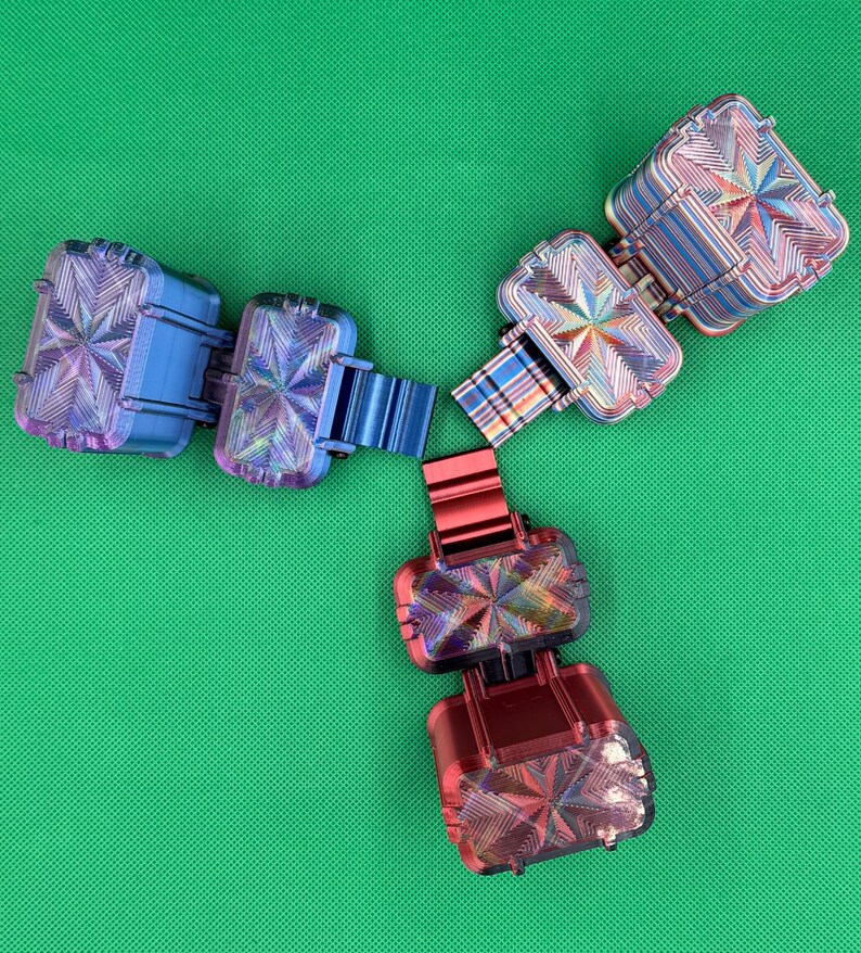Puede incluir: Tres juguetes antiestr&eacute;s articulados dispuestos en forma de Y. Cada juguete consta de dos bloques rectangulares conectados por una bisagra, con un acabado met&aacute;lico y un patr&oacute;n de estrella en la superficie. Uno es azul, otro rojo y otro iridiscente.