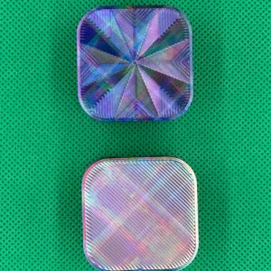 May include: Two square-shaped objects with rounded corners featuring iridescent, multi-colored surfaces. The top object displays a radial, star-like pattern of ridges, while the bottom object features a uniform, diagonal, linear texture.