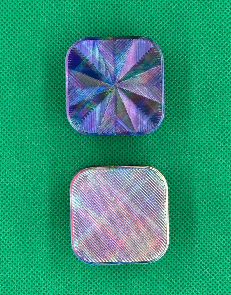 May include: Two square-shaped objects with rounded corners featuring iridescent, multi-colored surfaces. The top object displays a radial, star-like pattern of ridges, while the bottom object features a uniform, diagonal, linear texture.