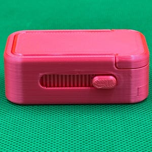 May include: A rectangular, hot pink 3D-printed storage box with rounded corners. The front side features a horizontal recessed track with a sliding button mechanism that moves along a series of vertical ridges, allowing for manual operation.