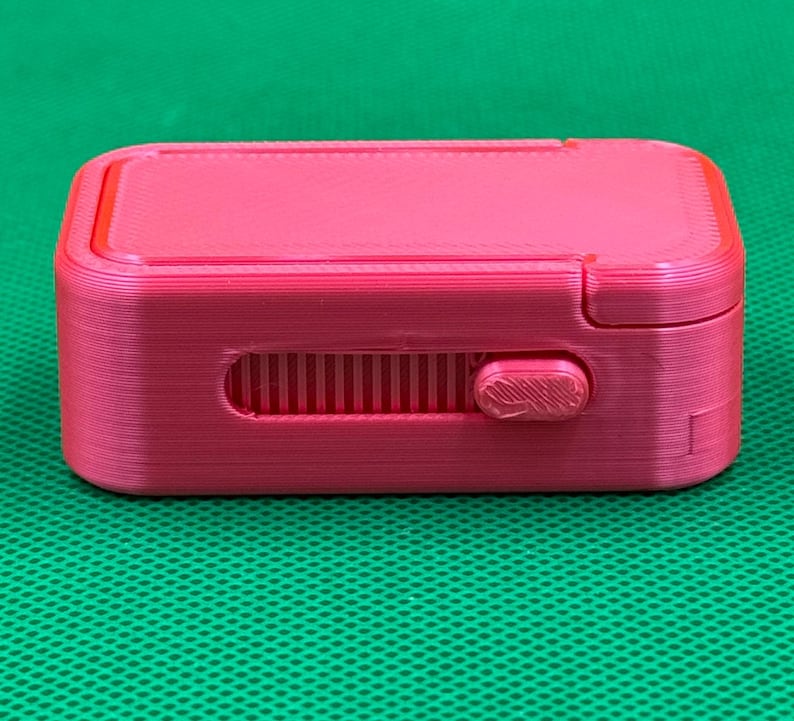 May include: A rectangular, hot pink 3D-printed storage box with rounded corners. The front side features a horizontal recessed track with a sliding button mechanism that moves along a series of vertical ridges, allowing for manual operation.