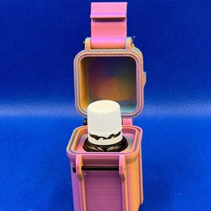 May include: A 3D-printed, pink and orange gradient storage case with a hinged lid, standing upright in an open position. Inside the square container sits a small, dark glass bottle with a white ribbed cap, securely held in place.
