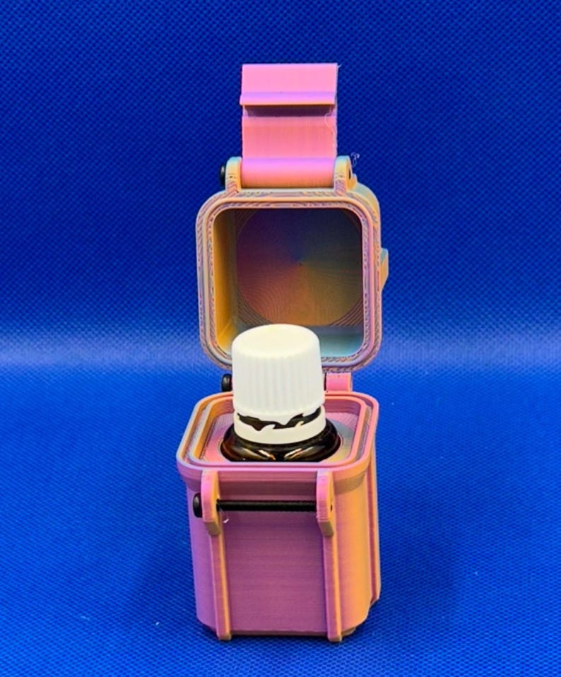 May include: A 3D-printed, pink and orange gradient storage case with a hinged lid, standing upright in an open position. Inside the square container sits a small, dark glass bottle with a white ribbed cap, securely held in place.
